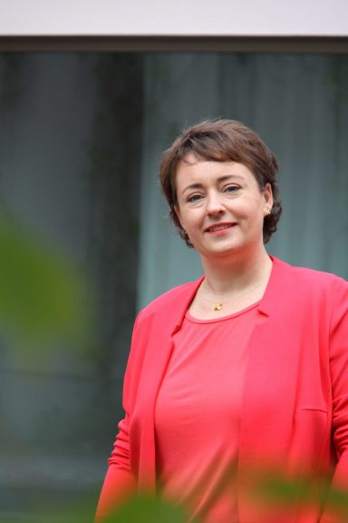 Frau mit kurzer brauner Haarfrisur, in einem roten Blazer, lächelt freundlich.