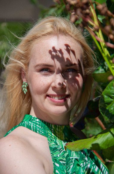 Junge Frau mit blondem Haar und grünen Kleid inmitten grüner Pflanzen.