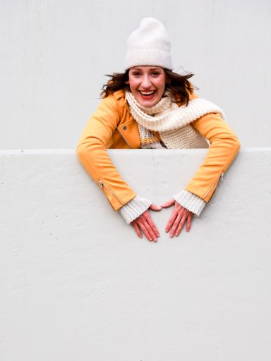 Lächelnde Frau in gelber Jacke und weißer Mütze, die an einer Wand lehnt.