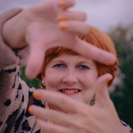 Frau mit rotblondem Haar, lächelt und zeigt mit den Fingern ein Quadrat.