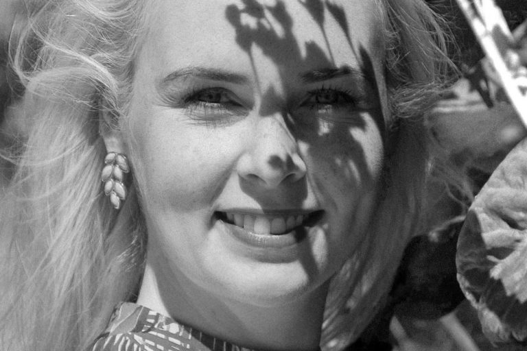Lächelnde Frau mit blonden Haaren, von Schatten einer Pflanze umgeben.
