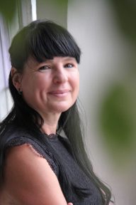 Lächelnde Frau mit langen, schwarzen Haaren, steht an einem Fenster.