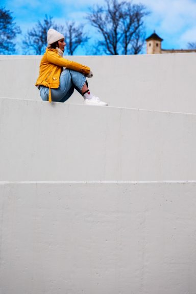 Junge Frau in einem gelben Pullover sitzt auf einer Mauer, blauem Himmel im Hintergrund.