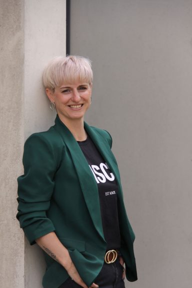 Blonde Frau im Businessoutfit steht vor einer hellen Wand.