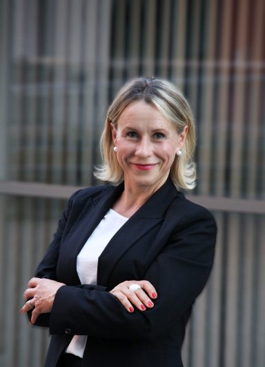 Professionelle Geschäftsfrau mit blonden Haaren, in einem schwarzen Anzug, lächelt freundlich.